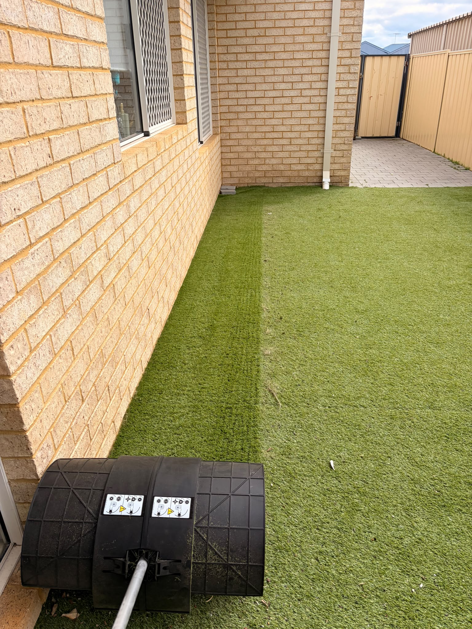 Artificial turf mid-clean, a worked strip of lifted, richer-green fibres next to the untouched, flatter turf beside it, STIHL KB-KM bristle brush visible in frame
